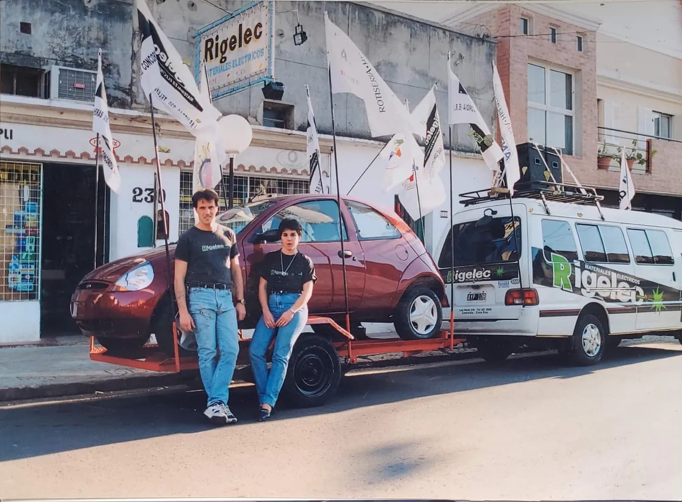 Dos integrantes del equipo junto a los autos y la camioneta de reparto, con banderines de Rigelec